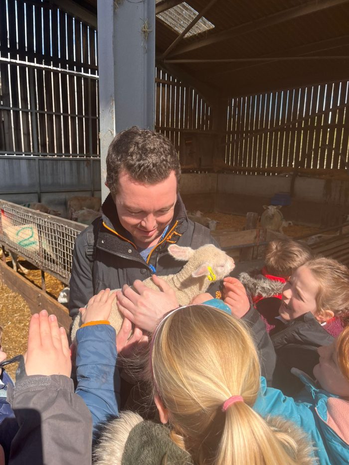A Teaching Assistant holds a lamb for the children at a farm.